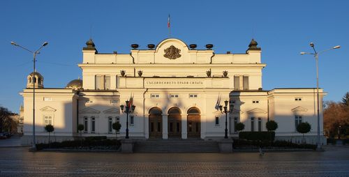 Българският парламент, наполовина огрян от сутрешното слънце, наполовина в сянка.