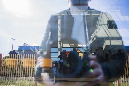 Отражение на фотографа в стъклата, през които се виждат бежанци, редящи се на опашка