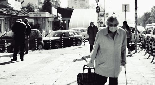 Хора на улицата, възрастна жена с маска, бастун и пазарска количка на преден план